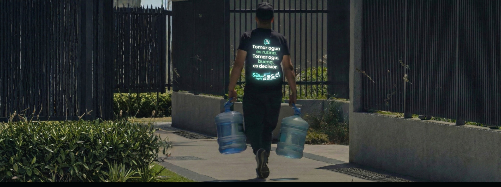 Repartidor de Sauces caminando con dos bidones de agua purificada premium para entrega a domicilio en sector residencial de Santiago.