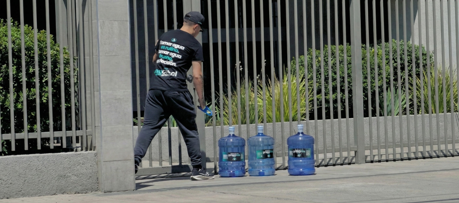 Repartidor de Sauces completando entrega de tres bidones de agua purificada en el acceso perimetral de un edificio residencial en Las Condes.